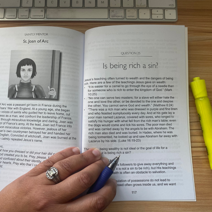 Open book with a question about being rich, held by a hand with a ring, on a wooden surface with a keyboard in the background.