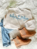 White sweatshirt with blue floral design, blue jeans, and brown boots on a textured surface.