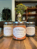 Three candles with labels on a wooden surface, with a blurred indoor background.