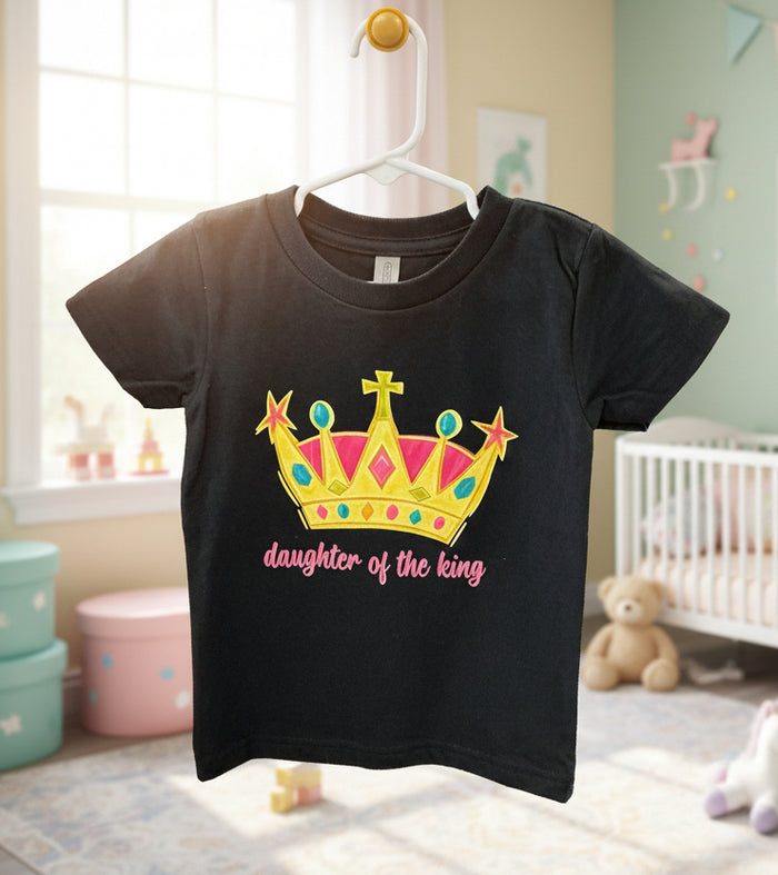 Black t-shirt with a colorful crown design and 'daughter of the king' text, hanging in a nursery.