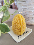 Decorative beeswax candle with floral patterns on a checkered cloth, next to a plant and woven basket.