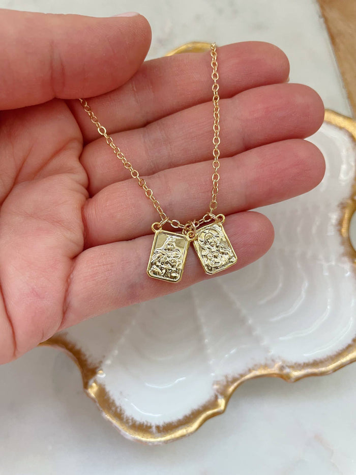 Gold necklace with two pendants held by a hand on a decorative plate.