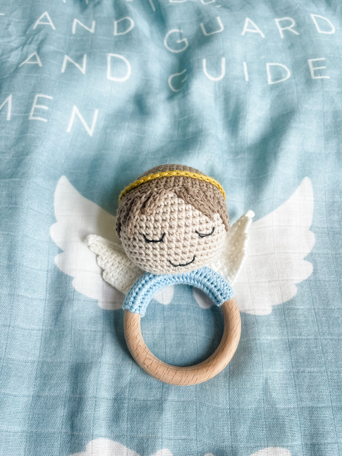 Crocheted angel rattle toy on a textured blue fabric background with embroidered text.