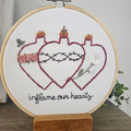 Embroidery hoop with a heart design and text 'inflame our hearts' on a white background.