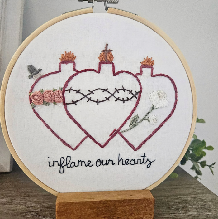 Embroidery hoop with a heart design and text 'inflame our hearts' on a white background.