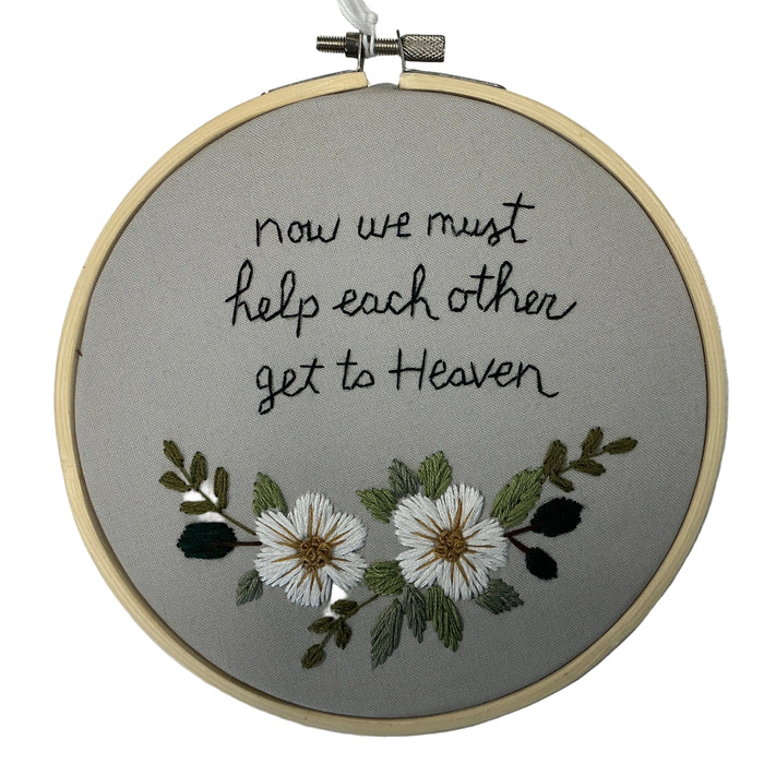 Embroidery hoop with text 'now we must help each other get to Heaven' and floral design on a gray background.