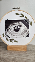 Embroidery hoop with ultrasound photo and green leaves on a wooden stand against a white wall.