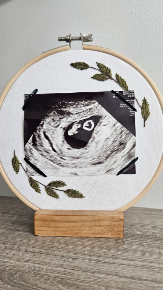 Embroidery hoop with ultrasound photo and green leaves on a wooden stand against a white wall.